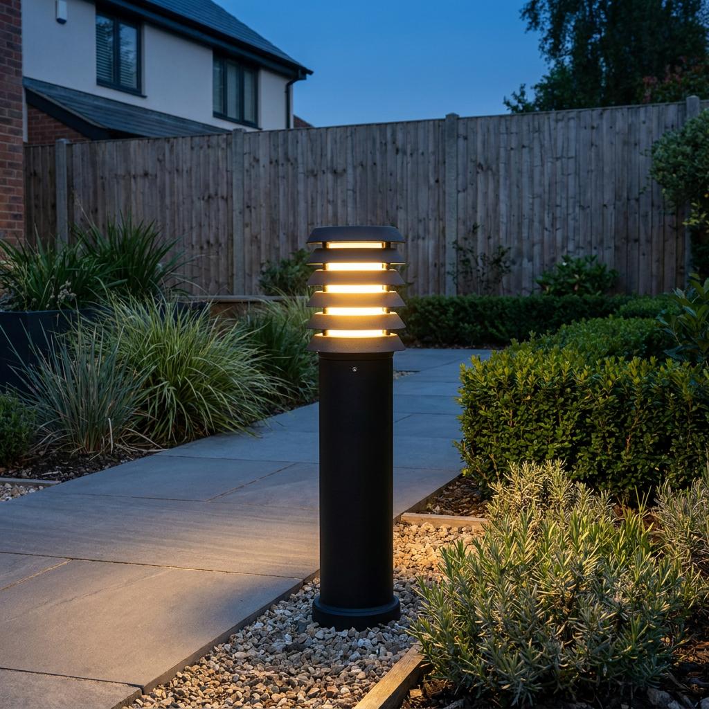 Elstead Alta mini black bollard light glowing warmly on a stone garden path at twilight.