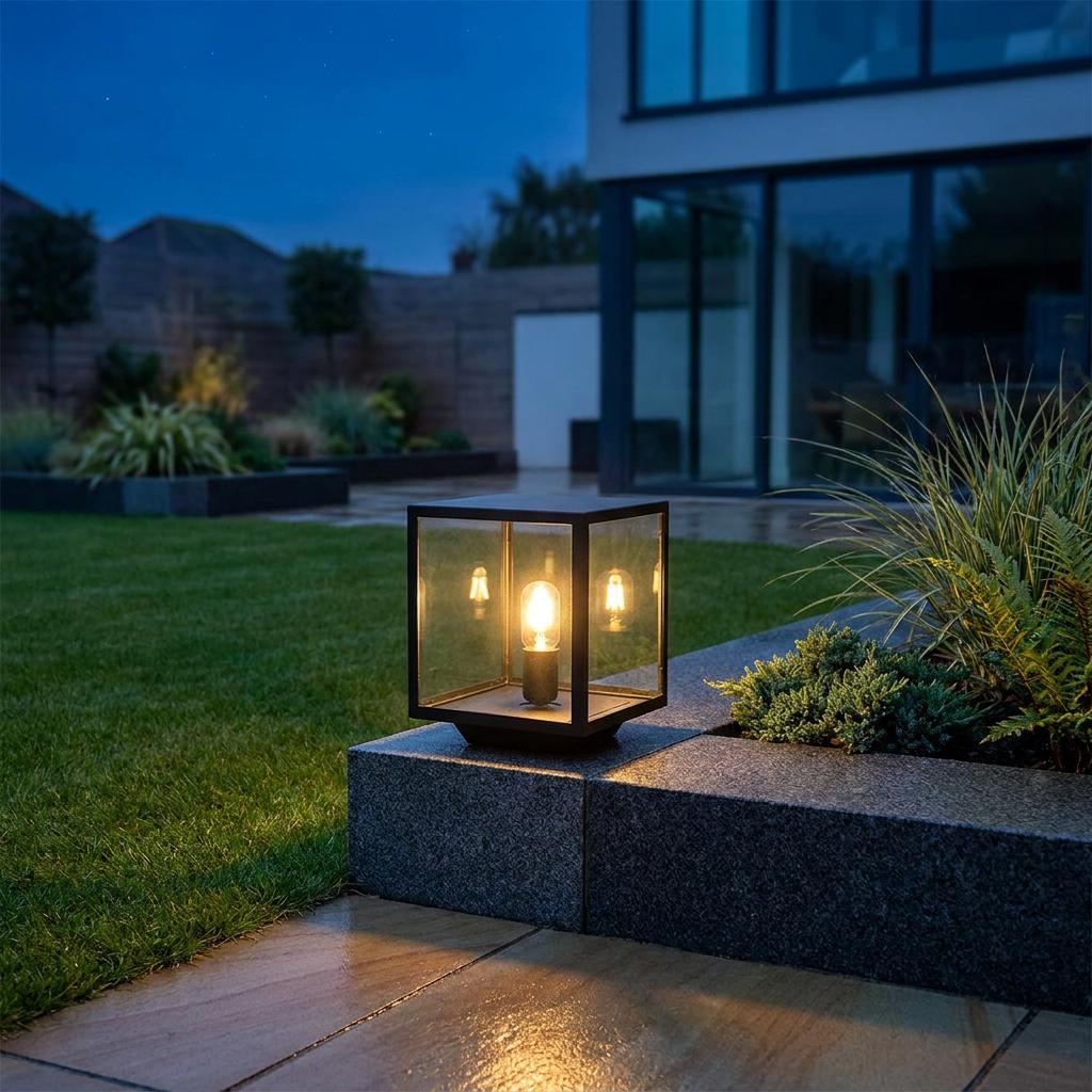 Black square pedestal lantern with a glowing bulb sits on a stone garden wall at dusk.