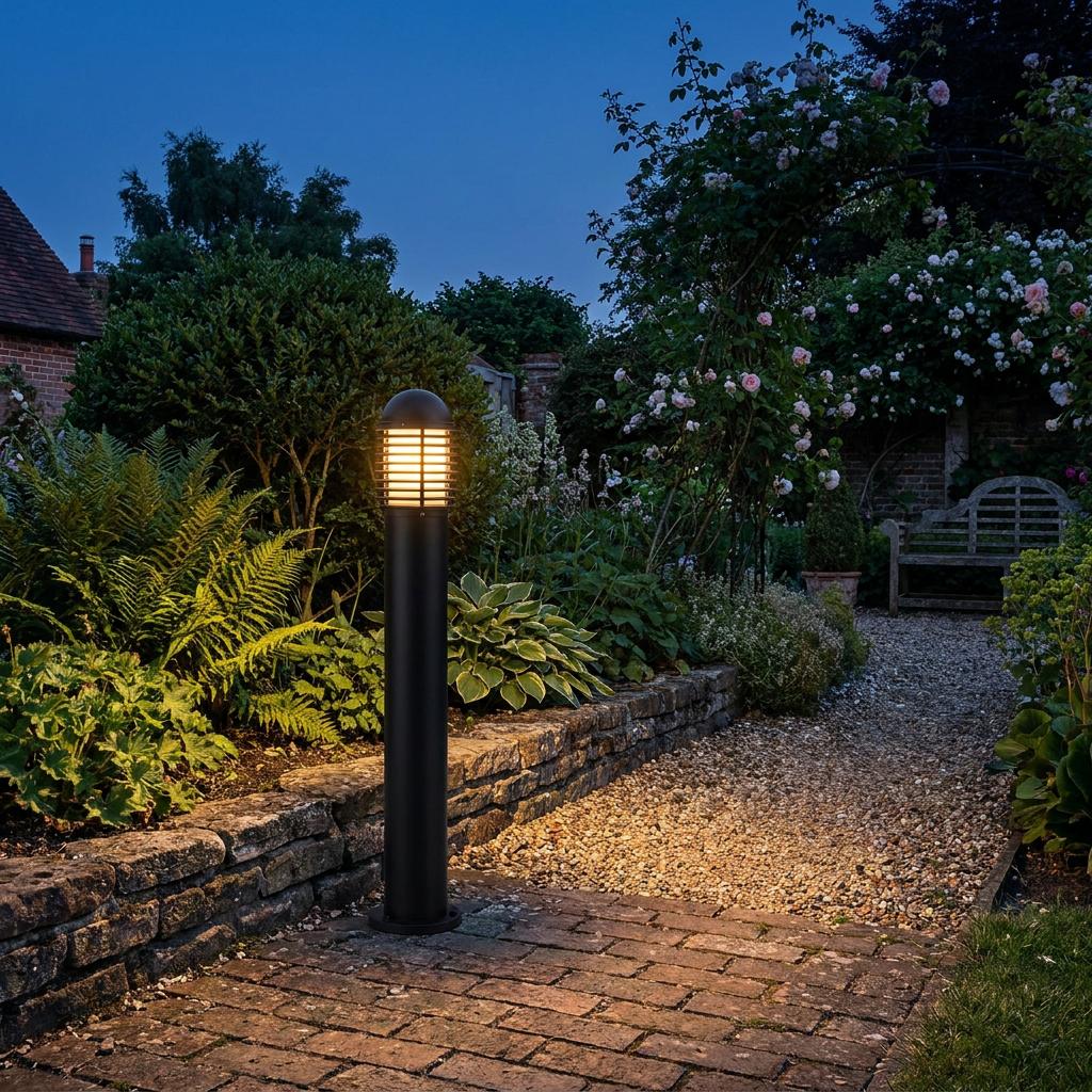 Matte black outdoor bollard light with a louvred top emitting a warm glow in a lush garden at dusk.