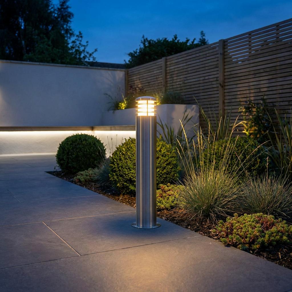 Illuminated stainless steel bollard light standing beside a garden path with shrubbery at twilight.