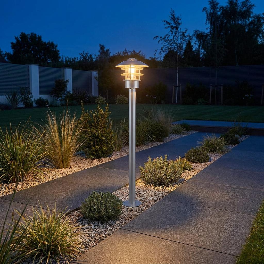 Silver Helsingor bollard lantern with a tiered shade glowing on a gravel garden path at twilight.