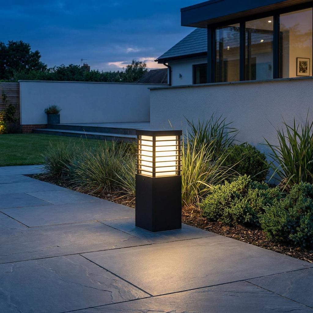 Black square bollard light with a slatted lantern top illuminating a modern patio path at dusk.