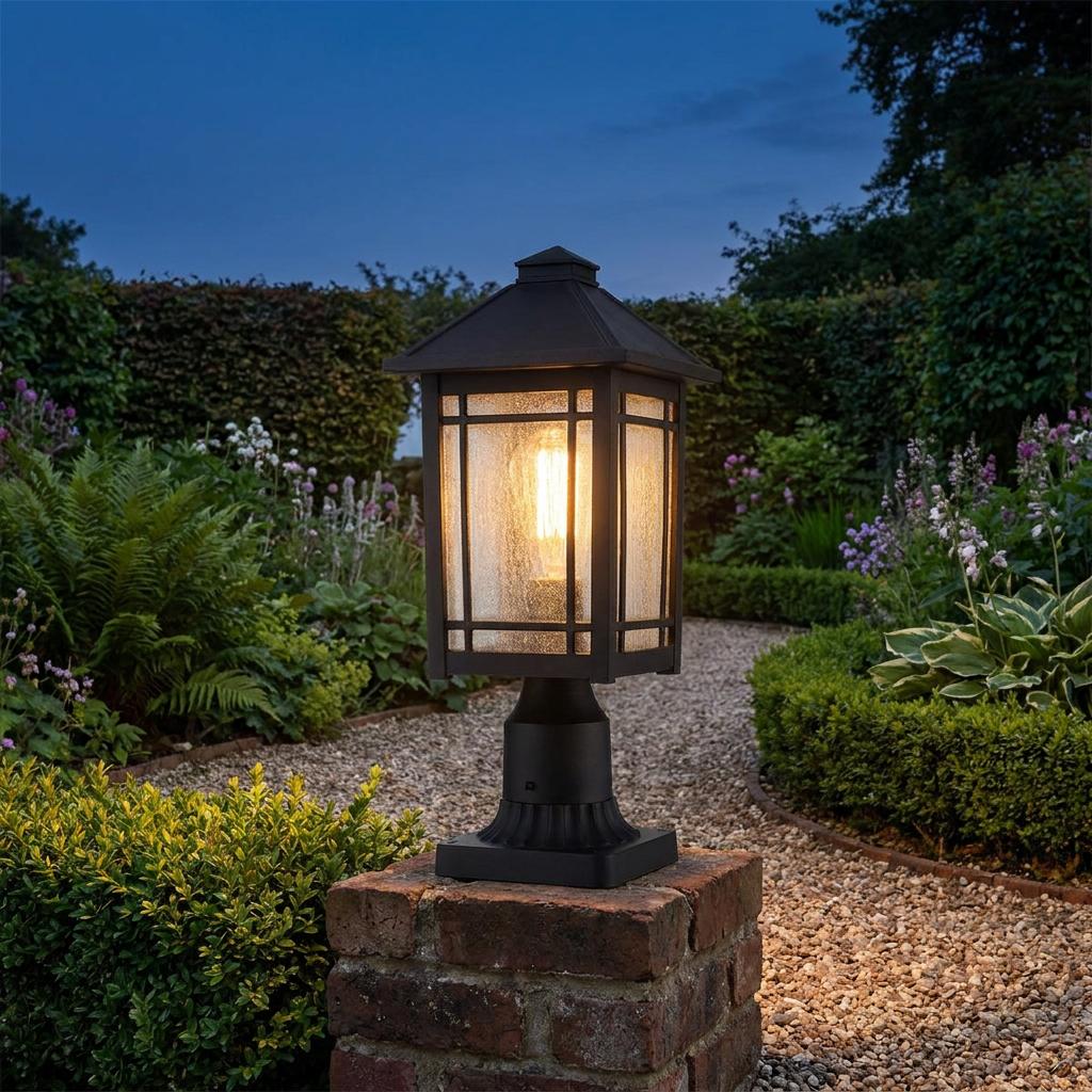 Black outdoor pedestal light with textured glass glowing on a brick post along a garden path at twilight.