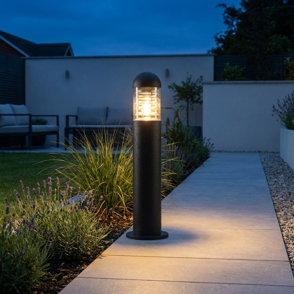 Black outdoor bollard light with a warm glow illuminates a modern garden path and nearby lavender at dusk.
